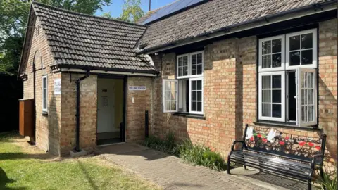 Kate Moser Andon/BBC A brick building with a pitched roof. A door is open and a sign reading "polling station" is hung to its left. A number of windows are open and a black bench is at its front, with is decorated with white doves and poppies.