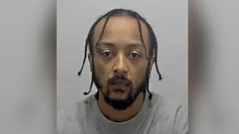 A mugshot of a man wearing grey with braised hair and a thick beard