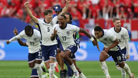 Getty Images Ivan Toney runs off in celebration with England teammates including John Stones, Jude Bellingham and Cole Palmer after the team beat Switzerland in a penalty shootout at Euro 2024. All are wearing white England shirts, with blue shorts and white socks.