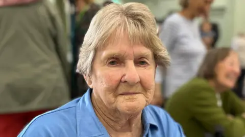 Tom Jackson/BBC Margret French has short blonde hair and is wearing a blue T-shirt. She is sitting down and is surrounded by several people.