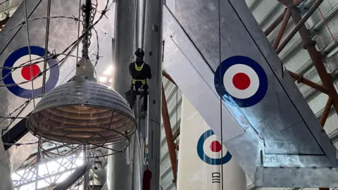 A person abseils down a silver RAF plane that is hanging from the ceiling of a hangar. 