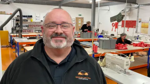 A man dressed in a black fleece with orange company branding in a large sewing workshop with women working at machines on workbenches behind him