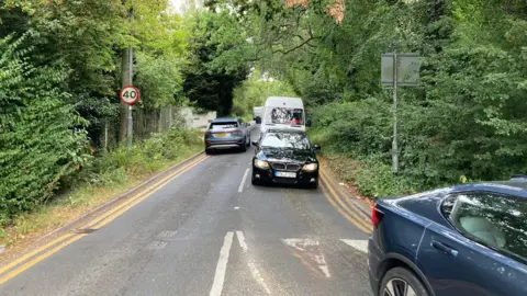 A number of cars and a white van driving up and down a busy single-carriageway road . There is a 40 miles per hour sign with a red ring and white background on one side of the road.