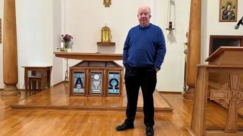 Fr Michael MacDonald is standing inside his church. He is wearing a blue jumper and black trousers and shoes.