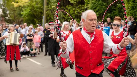 Electric Egg A man with short white hair in a red waistcoat, white shirt and red bandana around his neck is pulling a concentrated face as he danced with a red, black and white rope. Behind him is a woman playing a musical instrument and other dancers and spectators.