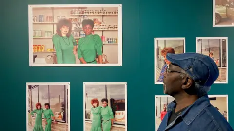 Charles Heslett/BBC A man looking at white-framed colour photographs hung up on a wall.