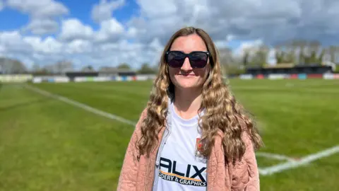 Tom Jackson/BBC Izzy Papandronicou smiling and looking at the camera. She is wearing large black sunglasses, a white Cambridge City FC jersey and a light pink jacket. Behind her is the club's pitch at Sawston. 
