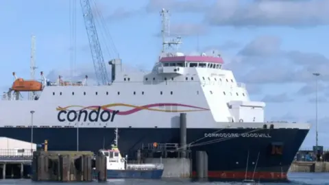 BBC A ferry with a blue hull and white topsides with the name Commodore Goodwill and Condor on the side berthed at a dock with a small boat in the foreground.