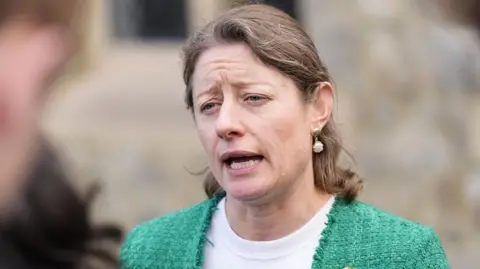 PA Media Helen Maguire, Liberal Democrat MP for Epsom and Ewell, speaks to the media outside Epsom Methodist Church in Ashley Road, Surrey. She is wearing a green jacket speaking to reporters outdoors, with a blurred figure in the foreground and a stone building in the background.