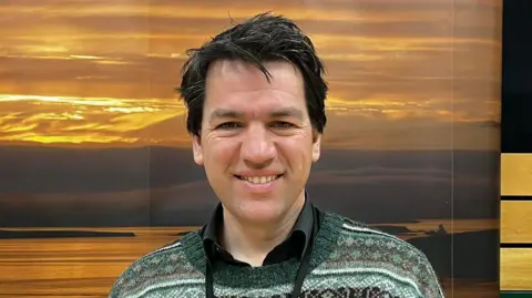 A man with dark hair and a patterned green and black jumper stands in front of a picture of a sunset