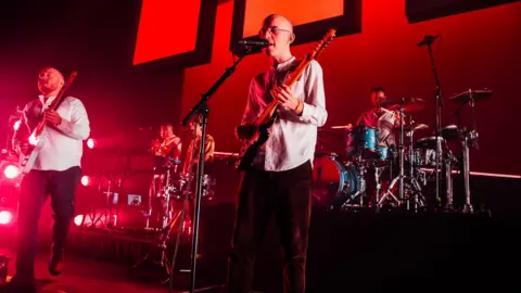 Getty Images Bombay Bicycle Club play on stage and there is a red hue over the stage. There is a guitarist playing and singing into a microphone in the middle of the stage.