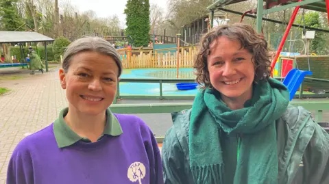 Sylwia Lipska is wearing a purple jumper and green collared T-shirt and Lucie Grange is wearing a green waterproof jacket and green scarf as they stand in front of the adventure playground. 
