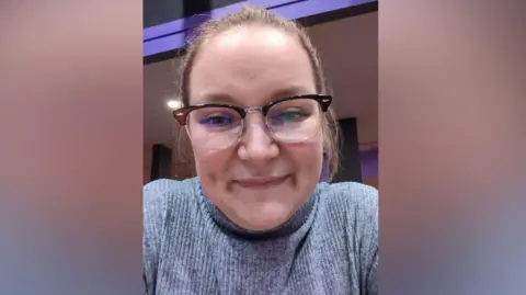 Micah Williams Micah wearing a grey, ribbed, long‑sleeved top while sitting indoors. A pair of dark-framed glasses is visible, and her hair appears to be tied back. The background includes softly lit interior features, with purple and warm-toned panels.