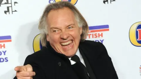 Getty Images Rik Mayall in front of a British Comedy Awards backdrop, laughing and pointing