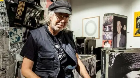 Ian Rook A man wearing a black cap, a black gilet and a black top is looking into the camera with a serious expression and wearing large clear-rimmed glasses. Posters and old photographs are all around him.