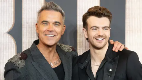 Getty Images Robbie Williams tersenyum lebar dan memeluk Jonno Davies yang juga tersenyum. Keduanya berpakaian rapi di acara perdana Better Man.