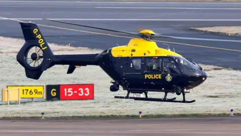 Roger Wasley A black and yellow Airbus H135 helicopter with police markings. It is hovering a few feet above the ground. The picture was taken at Birmingham Airport.