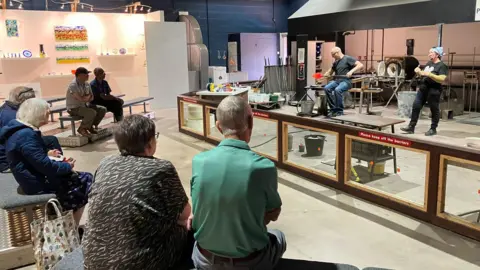 Six visitors can be seen sitting down on benches around a demonstration by two workers. One man is showing the glass being expanded while the other person gives a talk.