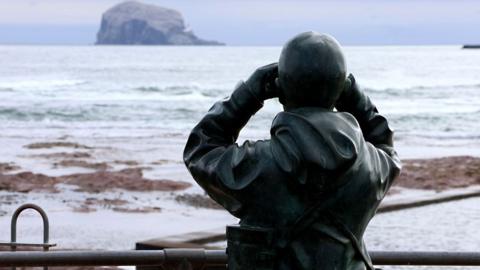 A statue of a man looking at the Bass Rock with binoculars