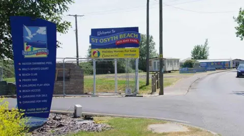 Blue signs at the entrance to St Osyth Holiday Park, which is based by the sea. A few static caravans are pictured in the background.