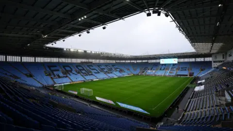 A photo from inside the stadium taken from high up and showing tiered blue seating. The pitch is empty and there is a large electronic screen high up at the far end behind one of the goals.