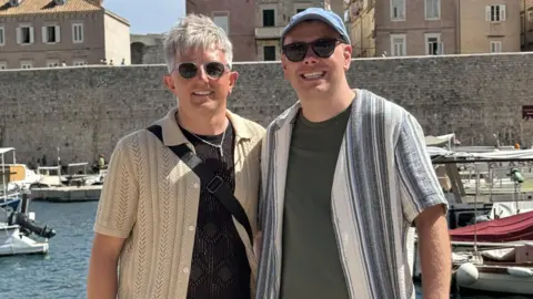Two men are stranding in front of a harbour with boats in the water. The man on the left has white blonde hair and is wearing sunglasses, a black top and beige shirt over the top. The man on the right is wearing a blue cap with a green/grey t-shirt and blue and white striped shirt on top.