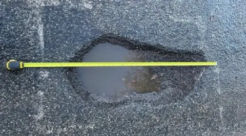 Jane Hodgson A deep pothole filled with water can be seen from above. A yellow tape measure is extended across the whole to demonstrate its size.