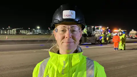 This is a head and shoulders picture of Brandi Davey. She is smiling and looking at the camera while wearing a hard hat with a sticker on it that says "Lagan". The background shows she is standing on a well lit runway at night. Over her left shoulder a multi storey car park can be seen. Over her right shoulder a number of construction vehicles can be seen and workers wearing high visibility clothing. 