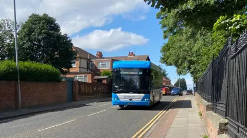 A blue and white Stagecoach bus driving along a road on sunny day with trees on both sides. There is a grey/red stone pavement on either side of the road, plus black metal railings on the right. In the distance, there are red-bricked residential houses.