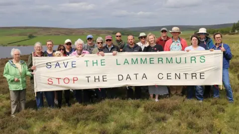 SLSDC A group of about 20 people standing behind a banner which says Save the Lammermuirs Stop the Data Centre. They are standing in rural countryside with a reservoir behind them.