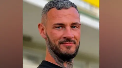 Ramon Logan has dark hair with grey streaks, a tattooed neck, dark beard and dark sweater