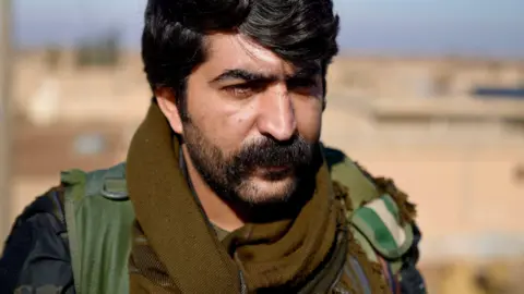 Goktay Koraltan/BBC Commander Azad of the Kurdish Forces stares past the camera. He wears a thick black moustache, heavy stubble, and his uniform. 