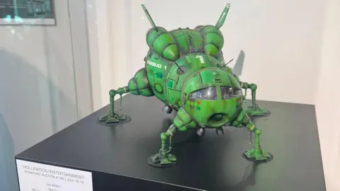 Heritage Auctions A model of Starbug 1, a ship from the series Red Dwarf. It is a green ship with four frog-like legs and a round cockpit. It is sitting on a plinth with a sign labelled "Hollywood Entertainment".