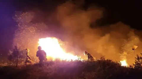Firefighters tackle a large blaze at night time. The billowing smoke is lit up orange and three firefighters are silhouetted against the large area of white flames in the centre of the picture.