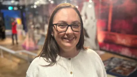 A woman in a white blouse smiles while standing inside in front of a display. She is wearing black glasses and has brown hair. 