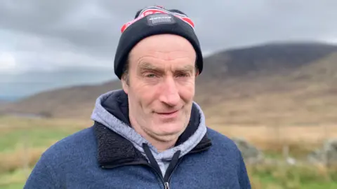 A man with a black and red woolly hat is smiling at the camera. He is wearing a grey hoody with a zip up navy fleece over it