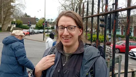 A smiling man with long hair in front of a metal fence.