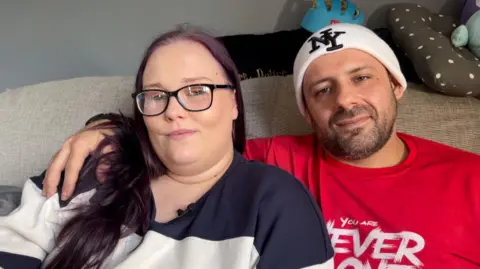 Angel-Kay and her husband Marley sitting on a sofa in their lounge. Angel is wearing a stripy top and Marley is wearing a red t-shirt and a white beanie hat.