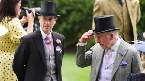 The King is holding the bill of his top hat and stood next to his brother, Prince Edward. Both are wearing formal suits.