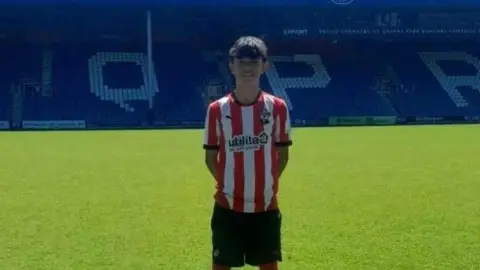 Chris McGeever Kai-Lloy is wearing a red and white striped football jersey, black shorts, and white socks stands on a green football pitch. In the background, there is a blue stadium stand with large white letters spelling “QPR” and signage reading “THE STANLEY BOWLES STAND.” The sky above is bright with scattered clouds.