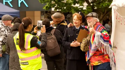 A woman in a hi-vis jacket takes a photo of a woman dressed in second world war RAF uniform and a man in a flat cap wearing a multi-coloured blanket