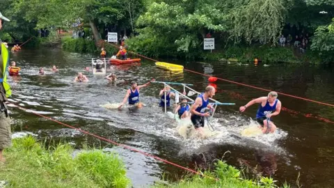 BBC/Oli Constable A team of runners wearing blue tops and black running shorts push a metal trolley between them, with one person sat in the middle through a river. Emergency services can be seen on standby in the water with safety equipment.