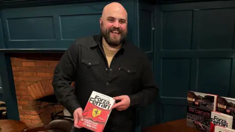 NATE SAUNDERS Nate Saunders smiles while holding a book with the title 'Forza Ferrari'. He appears to be standing in a pub or restaurant, with an emerald green painted wall behind him.