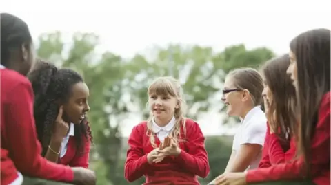 Five school children in red uniform sit around talking to each other with a backdrop of blurred trees. Stock image