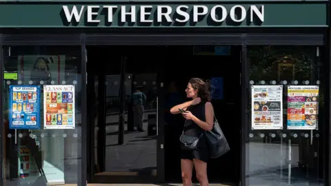 A woman dressed in a black vest and shorts stands outside a Wetherspoons holding her phone.