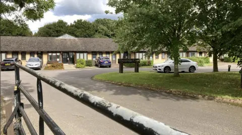 BBC An exterior view of the former care home at Wheatridge Court. There is a lone single story building with several cars parked outside and shrubs in front of the building