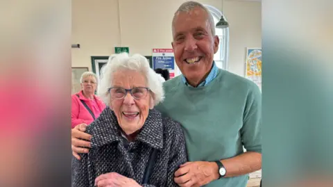 An elderly woman with glasses standing next to a man wearing a green jumper over a blue shirt. He has her arm around the woman. They are both smiling. 