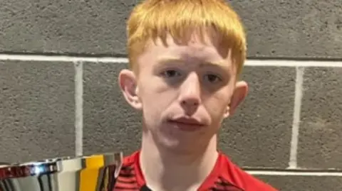 POLICE SCOTLAND Kory McCrimmon looking straight at the camera. He has light orange hair and is holding a trophy. He is wearing a red and black football shirt.