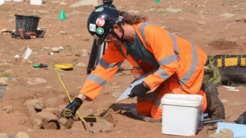 A man in orange high vis clothing and a black hard hat measures an area of brown soil on an excavation with a measuring tape