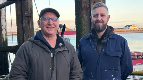 Tony and Phil Cowell standing by the sea. Tony is wearing a baseball hat and grey coat and has glasses. Phil is wearing dark blue overalls and has grey hair and a beard. Both are smiling
